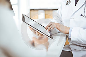 Unknown male doctor and patient woman discussing current health examination while sitting in clinic and using tablet