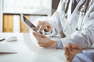 Unknown male doctor and patient woman discussing current health examination while sitting in clinic and using tablet