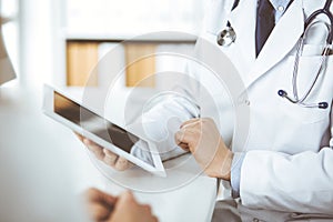 Unknown male doctor and patient woman discussing current health examination while sitting in clinic and using tablet