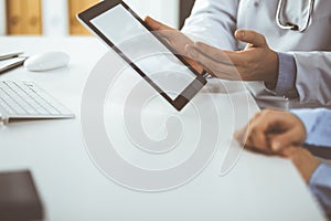 Unknown male doctor and patient woman discussing current health examination while sitting in clinic and using tablet