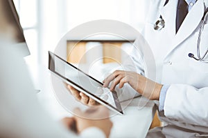 Unknown male doctor and patient woman discussing current health examination while sitting in clinic and using tablet