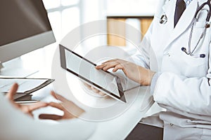 Unknown male doctor and patient woman discussing current health examination while sitting in clinic and using tablet