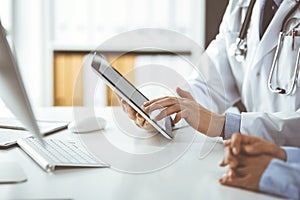 Unknown male doctor and patient woman discussing current health examination while sitting in clinic and using tablet