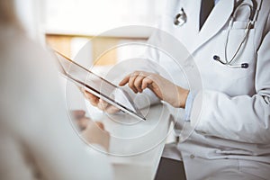 Unknown male doctor and patient woman discussing current health examination while sitting in sunny clinic and using