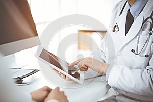 Unknown male doctor and patient woman discussing current health examination while sitting in sunny clinic and using