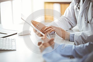 Unknown male doctor and patient woman discussing current health examination while sitting in sunny clinic and using