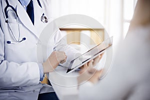 Unknown male doctor and patient woman discussing current health examination while sitting in sunny clinic and using