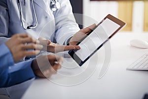 Unknown male doctor and patient woman discussing current health examination while sitting in sunny clinic and using