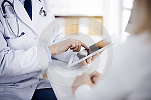 Unknown male doctor and patient woman discussing current health examination while sitting in sunny clinic and using