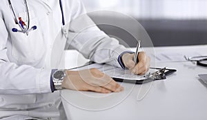 Unknown male doctor sitting and working with clipboard of medication history record in clinic at his working place