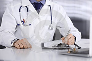 Unknown male doctor sitting and working with clipboard of medication history record in clinic at his working place