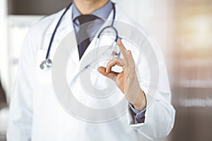 Unknown male doctor standing straight with OK sign in sunny clinic, closeup. Perfect medical service in hospital