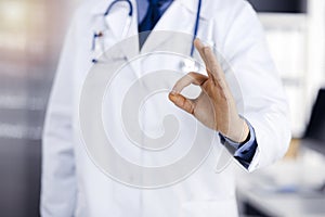 Unknown male doctor standing straight with OK sign in sunny clinic, closeup. Perfect medical service in hospital