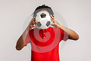 Unknown man with dreadlocks wearing red casual style T-shirt, hiding behind soccer ball, football fan covering his face during