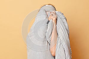 Unknown unrecognizable woman wrapped blanket and wearing sleeping eye mask isolated over beige background hiding her face do not