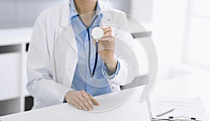 Unknown woman-doctor in blue blouse is holding stethoscope head while sitting at the desk in clinic, close-up. Physician