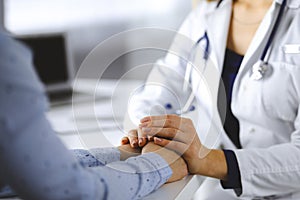 Unknown woman-doctor is holding her patient`s hands, discussing current health examination, while they are sitting at