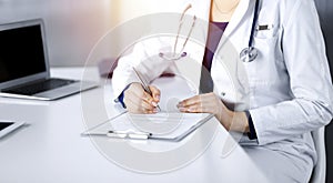 Unknown woman-doctor is prescribing some medication for her patient, using a clipboard, while sitting at the desk in her