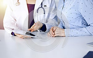 Unknown woman-doctor is showing to her patient a description of medication, while sitting together at the desk in the