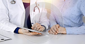 Unknown woman-doctor is showing to her patient a description of medication, while sitting together at the desk in the