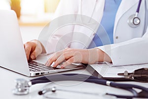 Unknown woman-doctor typing on laptop computer while sitting at the table in sunny clinic