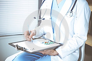 Unknown woman doctor at work at hospital. Pills at medical clipboard. Young female physician write prescription or