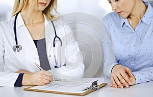 Unknown woman-doctor is writing down on her papers patient`s symptoms of a cold, while sitting together at the desk in