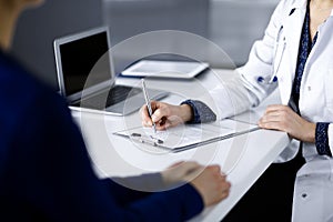 Unknown woman-doctor is writing some medical recommendations to her patient, while they are sitting together at the desk