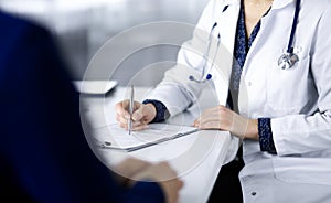 Unknown woman-doctor is writing some medical recommendations to her patient, while they are sitting together at the desk