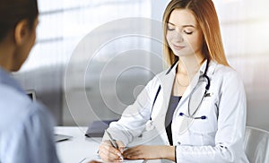 Unknown woman-doctor is writing some medical recommendations to her patient, while they are sitting together at the desk