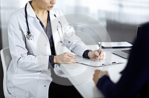 Unknown woman-doctor is writing some medical recommendations to her patient, while they are sitting together at the desk