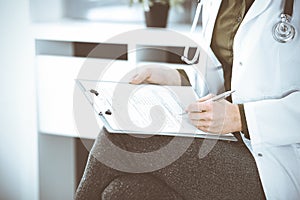 Unknown woman-doctor writing something at clipboard while sitting at the chair, close-up. Therapist wearing green blouse