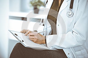 Unknown woman-doctor writing something at clipboard while sitting at the chair, close-up. Therapist wearing green blouse