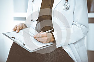 Unknown woman-doctor writing something at clipboard while sitting at the chair, close-up. Therapist wearing green blouse