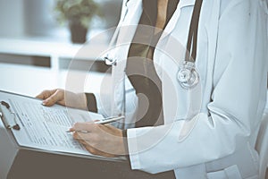 Unknown woman-doctor writing something at clipboard while sitting at the chair, close-up. Therapist wearing green blouse