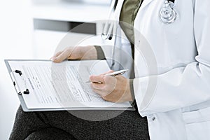 Unknown woman-doctor writing something at clipboard while sitting at the chair, close-up. Therapist wearing green blouse