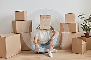 Unknown woman wearing white T-shirt sitting on the floor near cardboard boxes, posing with carton box on her head with drawing