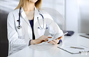 Unknown young woman-doctor is checking some medication names, while sitting at the desk in her cabinet in a clinic