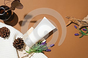 An unlabeled cosmetic tube, fresh lavender flowers, a brown glass vase, dried branches, pine cones and white fabric on a brown