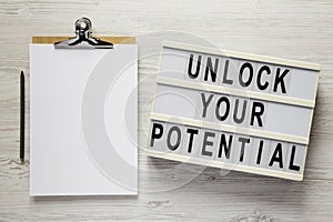 `Unlock your potential` on a lightbox, clipboard with blank sheet of paper on a white wooden background, top view. Flat lay,