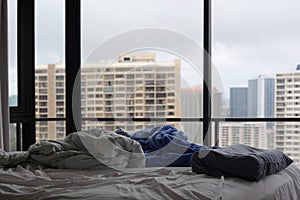 Unmade bed with messy blankets and pillow in hi-rise apartment with city view