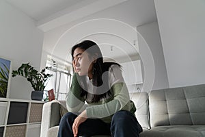 Unmotivated pensive Asian girl student age sits on couch in apartment leaning head on hand