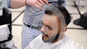 Unparalleled barber with a beard and a tattoo is cutting the hair of his client in the barbershop.