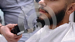 Unparalleled barber with a beard and a tattoo is cutting the hair of his client in the barbershop.