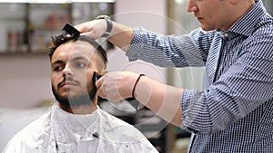Unparalleled barber with a beard and a tattoo is cutting the hair of his client in the barbershop.