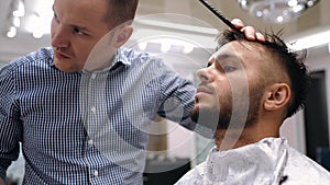 Unparalleled barber with a beard and a tattoo is cutting the hair of his client in the barbershop