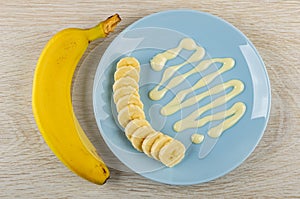 Unpeeled banana, slices of banana, condensed milk in plate on table. Top view