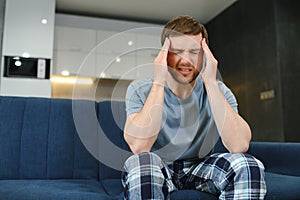 Unpleasant pain. Sad unhappy handsome man sitting on the sofa and holding his forehead while having headache.