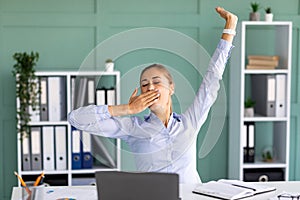 Unproductive worker. Young lady yawning and stretching hands while sitting at workplace in modern office