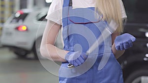 Unrecognisable blond woman in blue work robe and gloves holding wrench. Young female auto mechanic standing at the